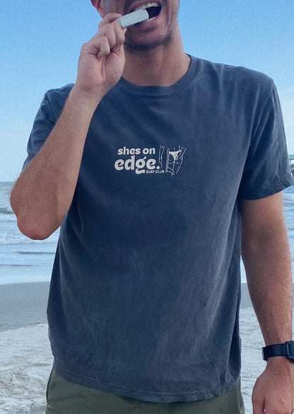 A young man bites a bar of surf wax while posing on a beach. He is wearing a Large Pepper SHES ON EDGE t-shirt. The image on the shirt is of a woman in a bikini holding a surfboard while picking a wedgie. The text says SHES ON EDGE surf club. The whole graphic is cream.