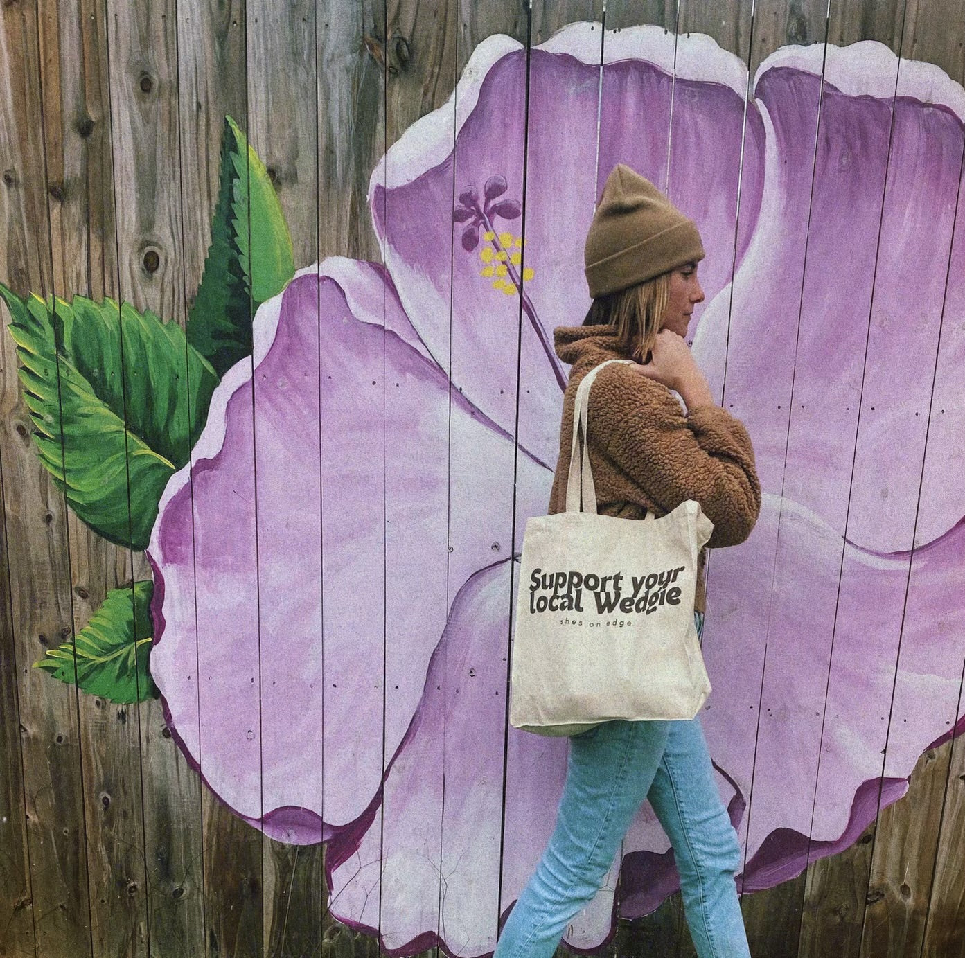 This image depicts a woman in a taupe beanie and jacket carrying a cream tote bag with brown writing that says "Support your local Wedgie shes on edge." The bag is being walked in front of a mural of a purple tropical flower. The woman is wearing jeans and has short, dirty-brown hair.