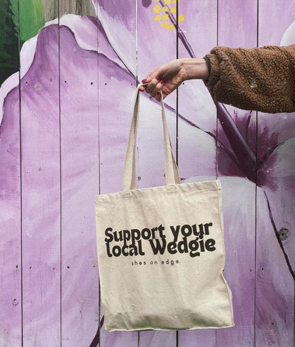 This image depicts a cream tote bag with brown writing that says "Support your local Wedgie shes on edge." The bag is being held in front of a mural of a purple tropical flower.  