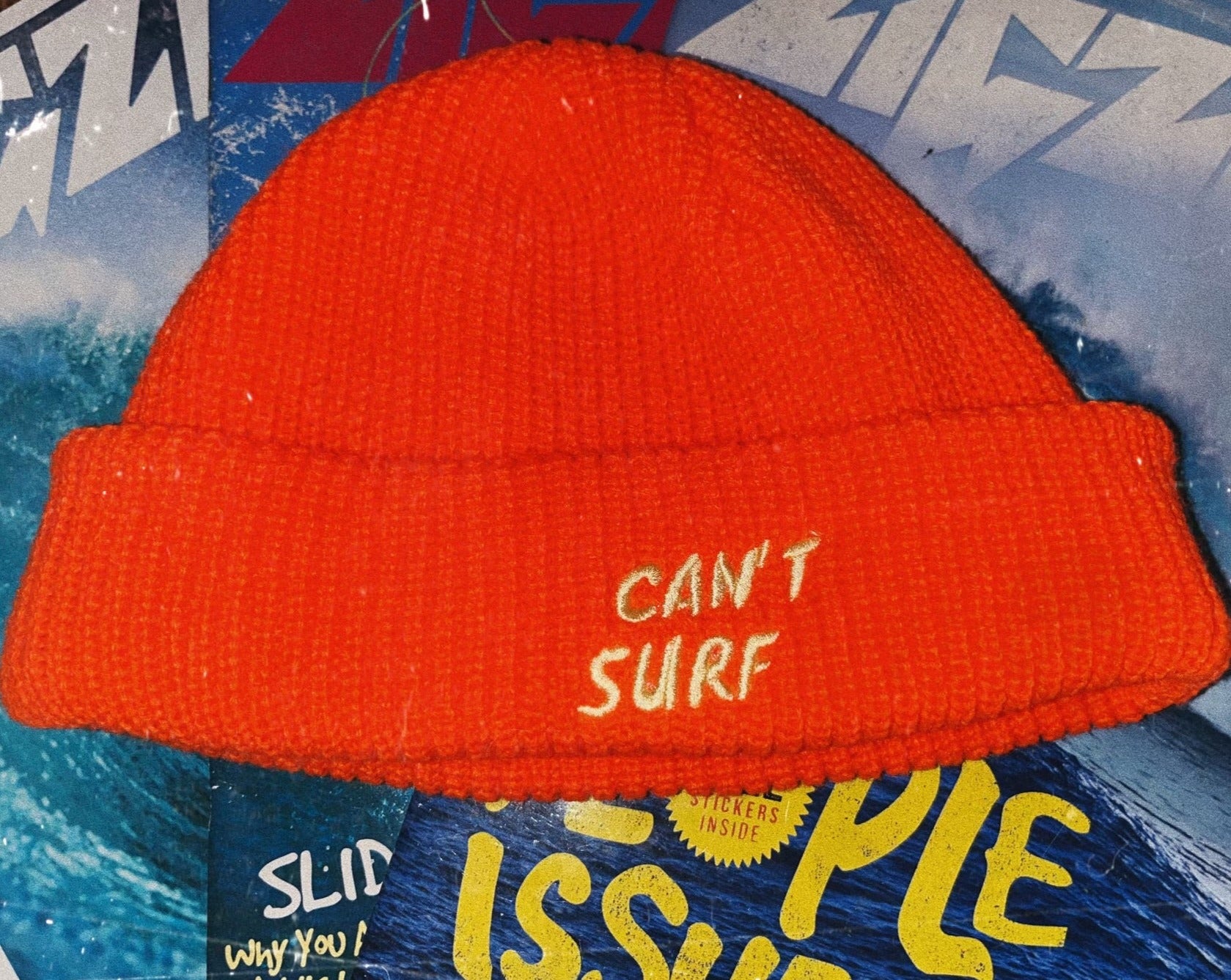 This image features a bright orange beanie that says "CAN'T SURF" in light yellow. The beanie is ribbed and is a fisherman's style cap. The hat is resting on top of a few surf magazines.