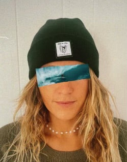 This photo features a woman with long, dirty-blonde hair wearing a black-colored SHES ON EDGE beanie. The beanie has a tag on the front featuring the Sally logo along with the text "shes on edge a womxn's surf collective". The woman's eyes are covered by a photo of a wave.