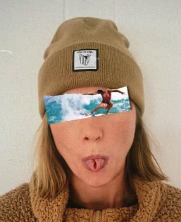 This photo features a woman with short, dirty-brown hair wearing a sand-colored SHES ON EDGE beanie. The beanie has a tag on the front featuring the Sally logo along with the text "shes on edge a womxn's surf collective". The woman is sticking out her tongue and her eyes are covered by a photo of a woman surfing. 